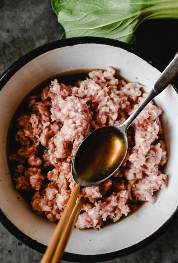 Top view of uncooked minced pork in a bowl with bok choy and spoon for food preparation.